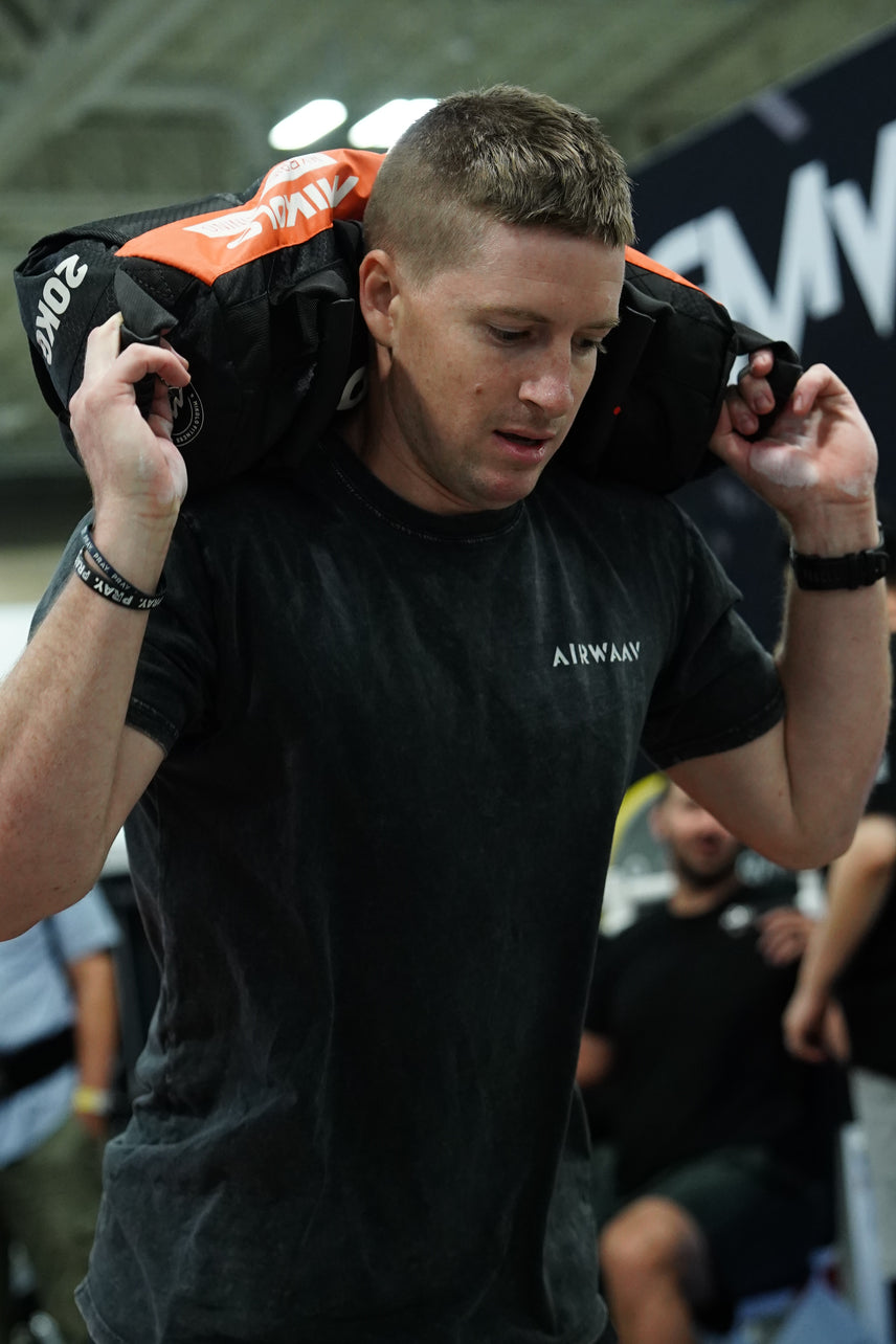 Man in a black t-shirt carrying a heavy 20kg black and orange sandbag on his shoulders.