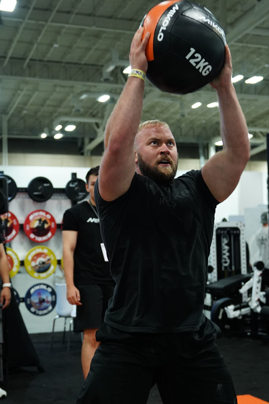 Bearded man in a black shirt lifting a 12kg black and orange Mikolo medicine ball overhead with both arms extended