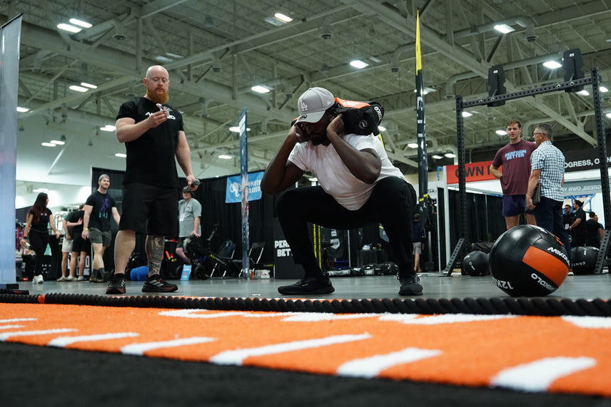 Man in a grey cap performs a deep squat holding a weighted bag on his shoulders while a trainer watches