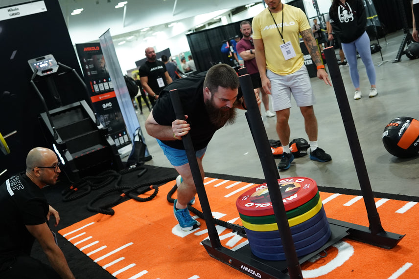 A bearded man pushes a MIKOLO sled loaded with colorful weight plates across an orange mat while a kneeling man watches nearb