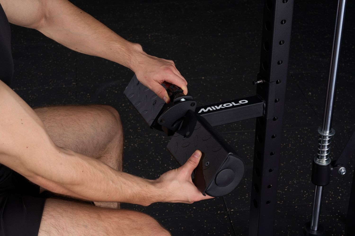 A person adjusts the angle of a black Mikolo lat pulldown and low row pad attachment on a gym rack