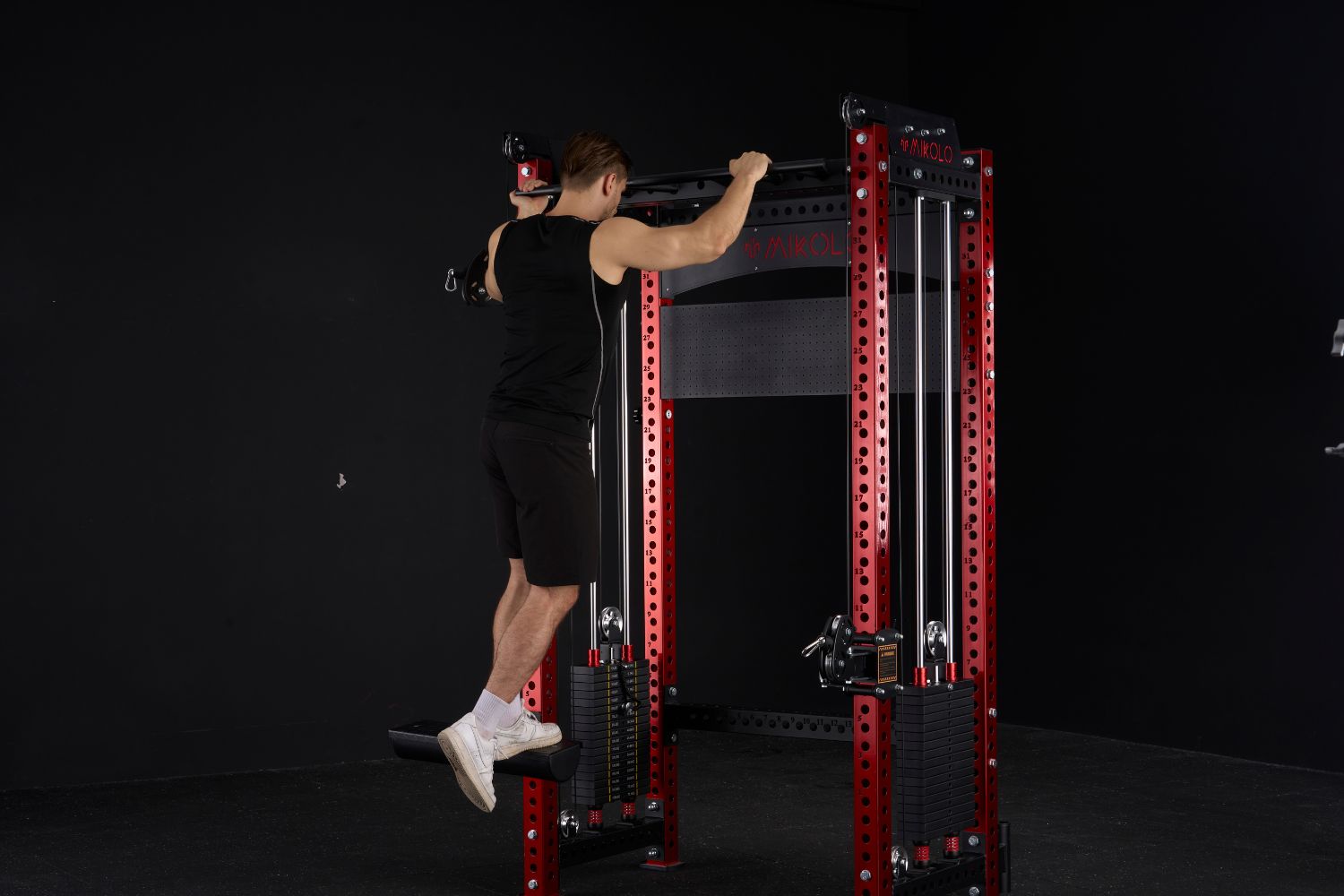 Athletic man in black uses a red Mikolo power rack, holding the top bar with feet resting on a black low row pad attachment