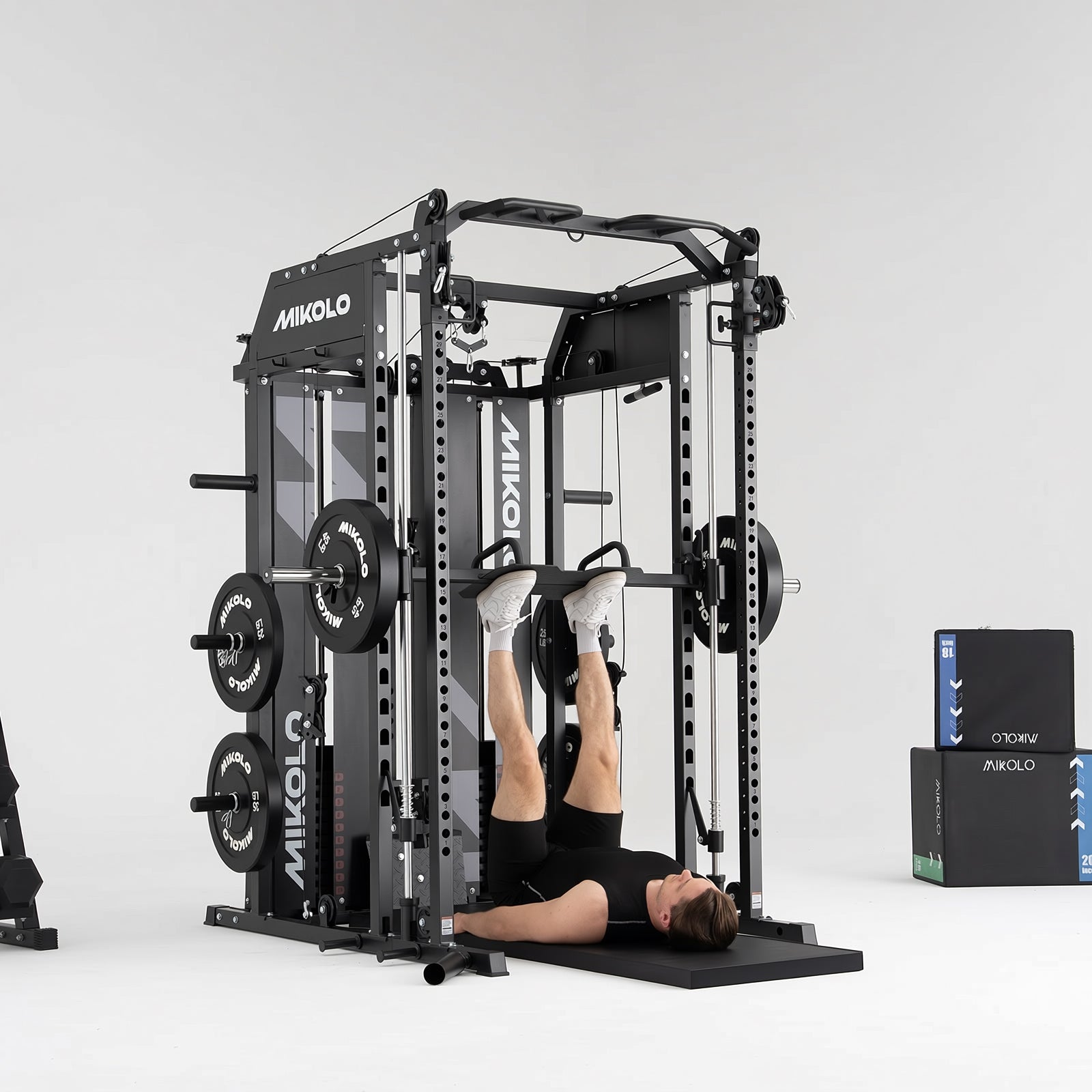 Man lying on back performing leg press on a black Mikolo Smith machine and functional trainer with weight plates