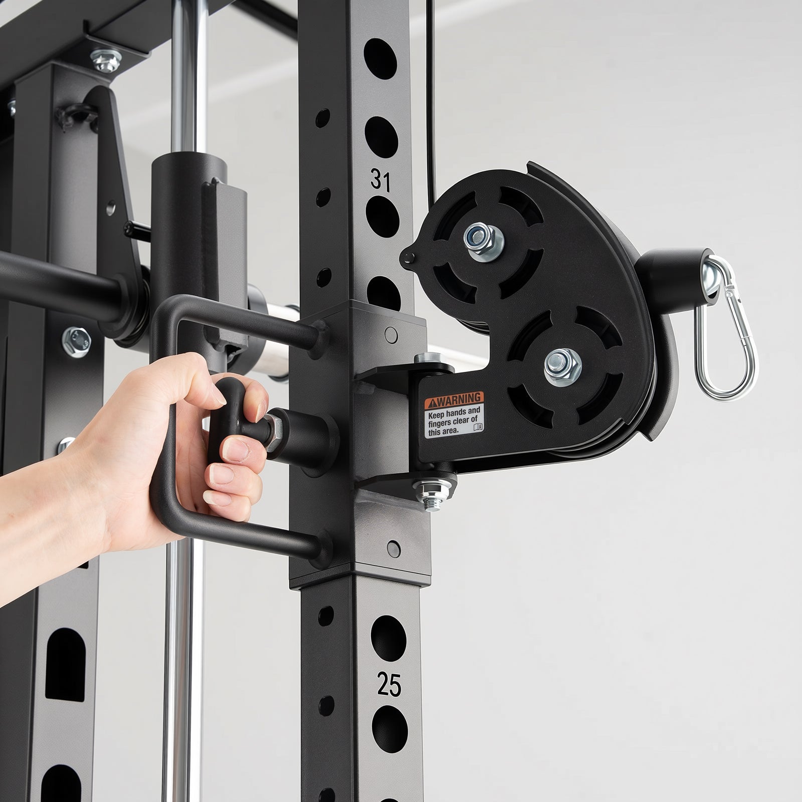 A hand adjusts a handle on a black metal power rack frame next to a dual pulley cable attachment with a silver carabiner