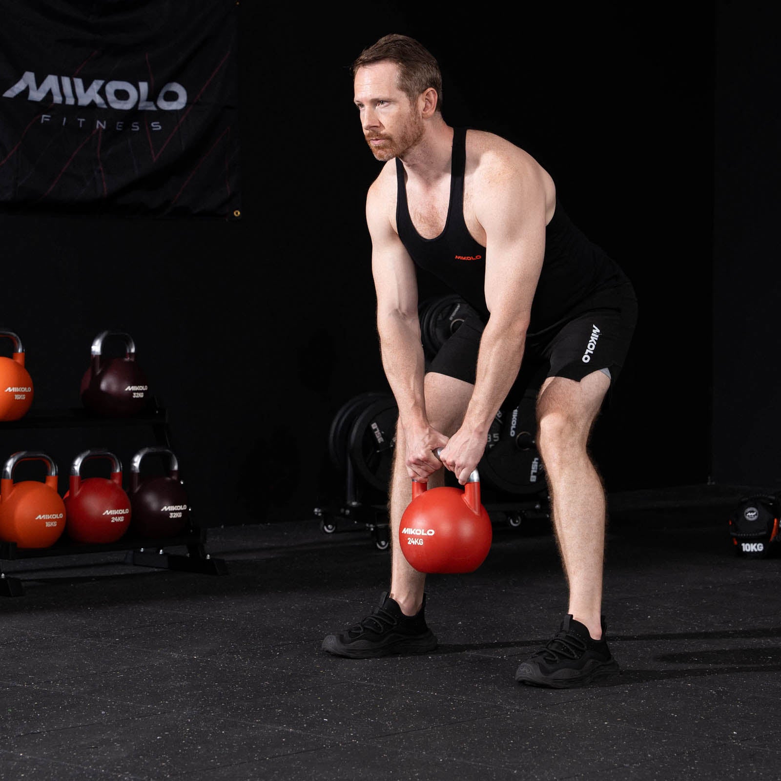 Man in black athletic wear holding a red 24kg Mikolo kettlebell with both hands between his legs in a gym setting