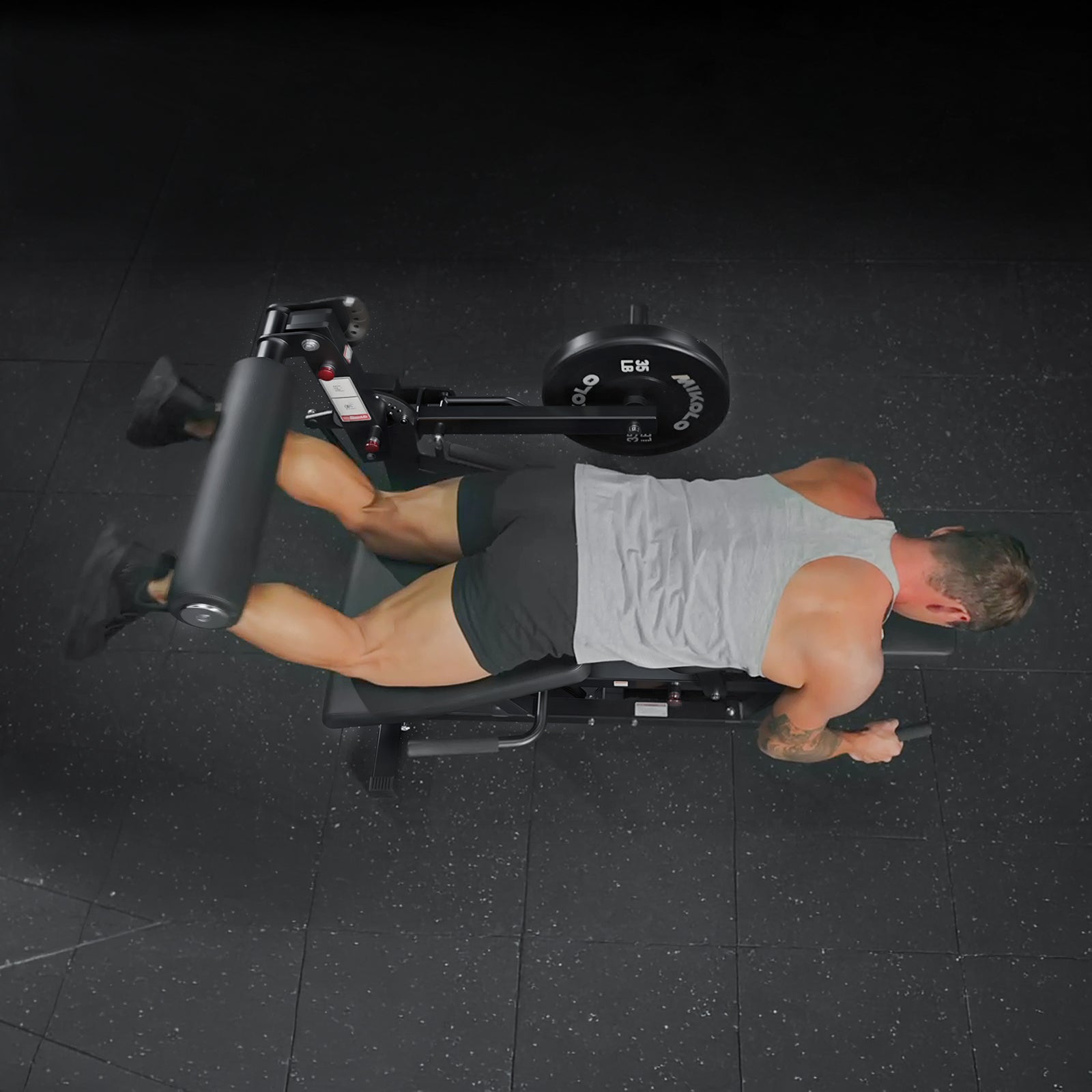 A man lies face down on a prone leg curl machine, bending his knees against a padded roller to lift a 35 lb weight plate