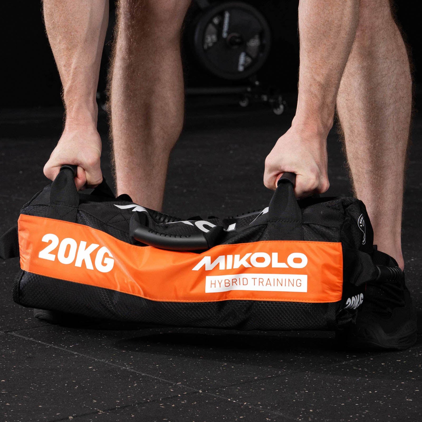 A person gripping the handles of a black and orange Mikolo 20kg hybrid training sandbag on a gym floor