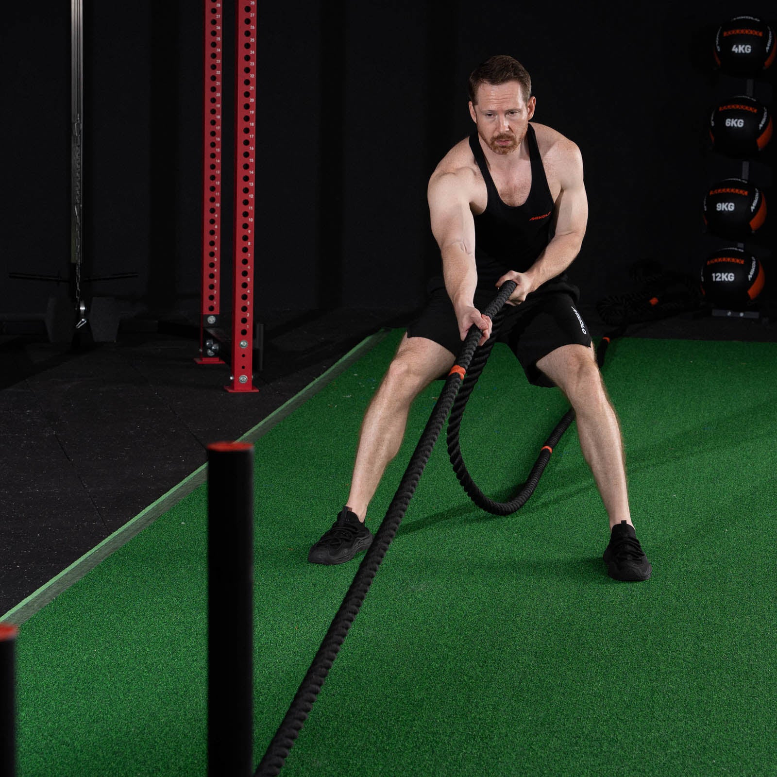 A muscular man in a black tank top and shorts pulls a thick black rope on green turf in a gym
