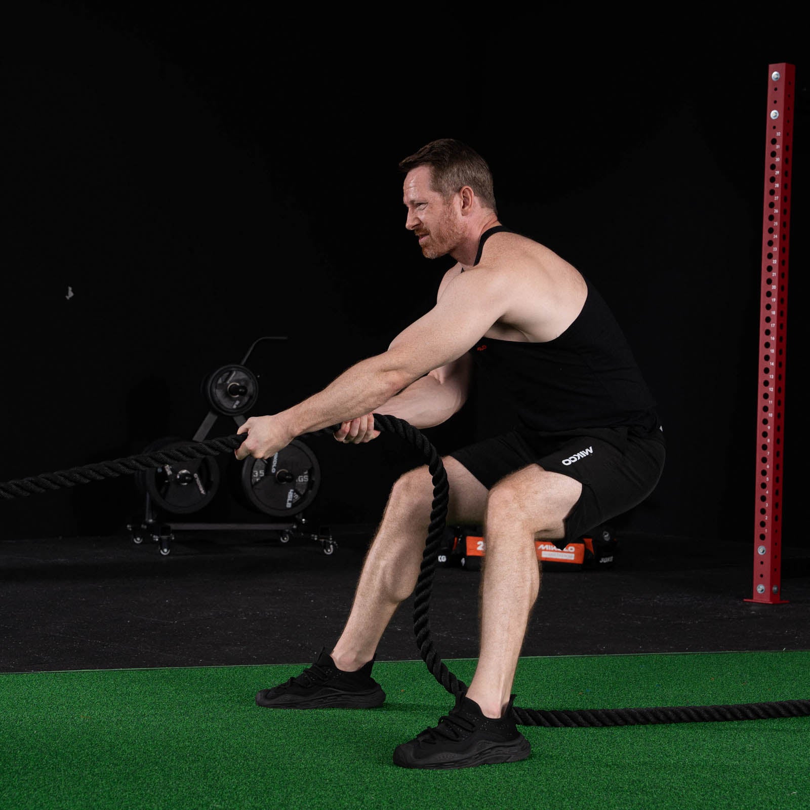 A man in a black tank top and shorts squats on green gym turf while pulling a thick black rope