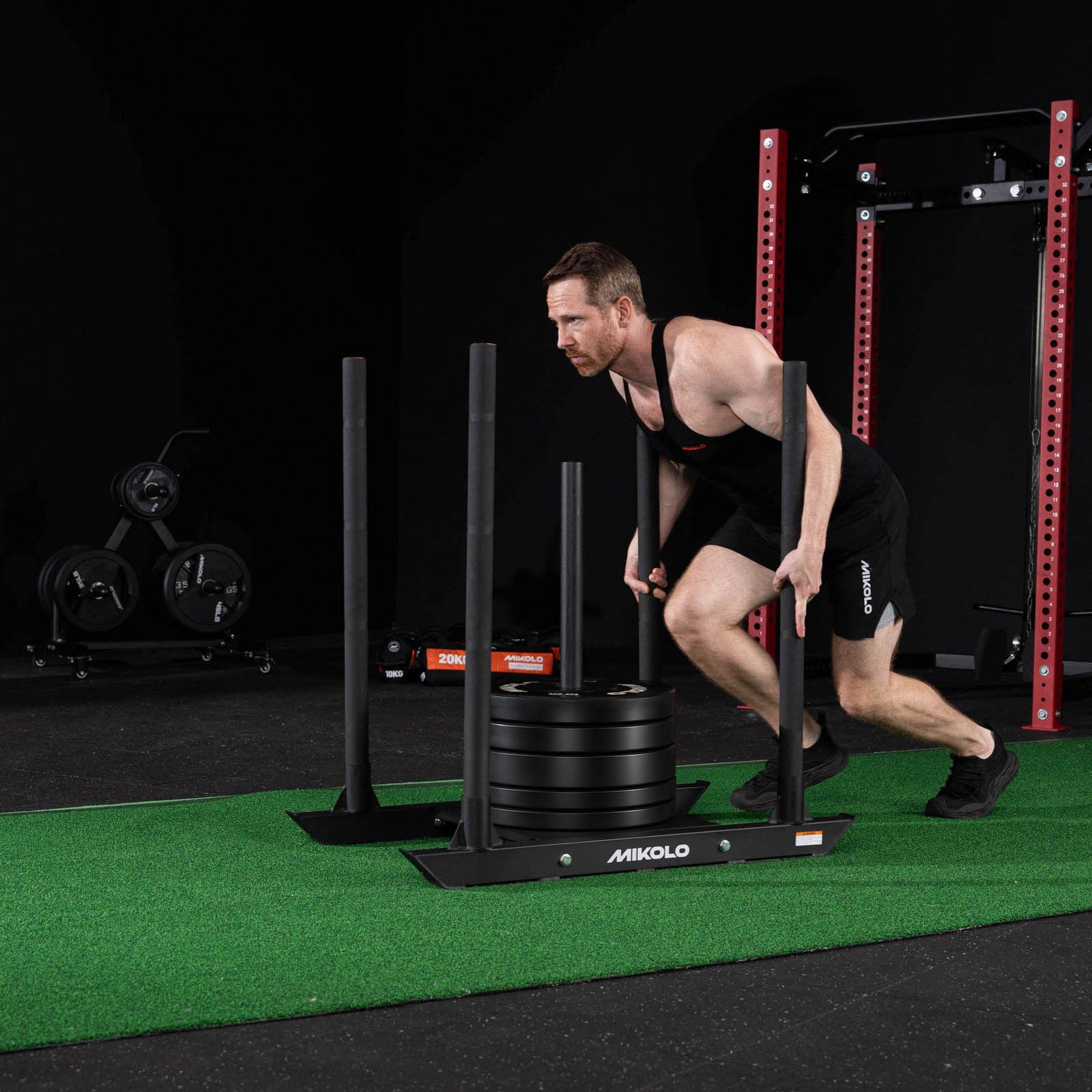 A man pushes a black MIKOLO weighted sled loaded with plates across green turf in a gym
