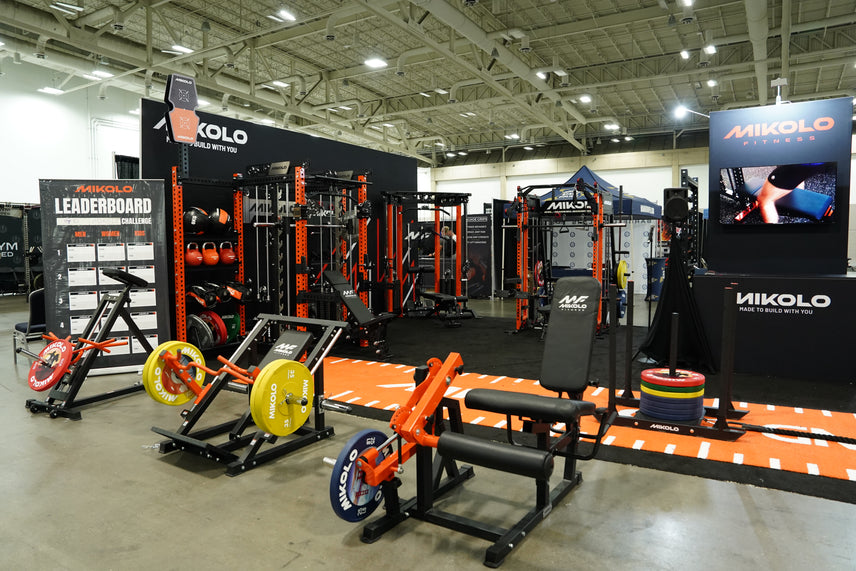 Mikolo Fitness booth featuring black and orange squat racks, benches, and plate-loaded machines with yellow and blue weight p