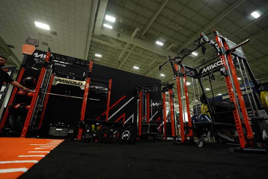 Red and black MIKOLO power racks and cable machines stand on black flooring beside orange turf with weight plates nearby