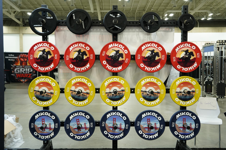 Rack of Mikolo plates: top row black 10-45 lbs, then red, yellow, and blue 25 lb plates with cowboy, beach, and bridge art