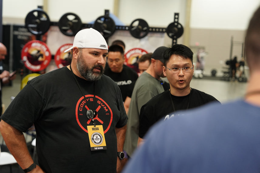 A bearded man in a white cap and media badge talks to a man with glasses in a gym with weight racks in the background