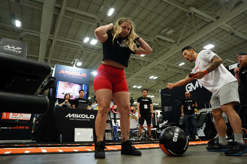 A woman in red shorts stands near a 6kg medicine ball while a man gestures towards her at a Mikolo Fitness booth