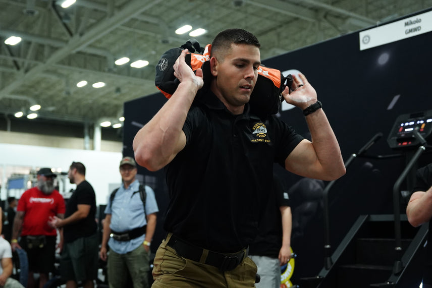 A man wearing a black Kentucky State Police Academy Staff shirt carries a weighted sandbag on his shoulders