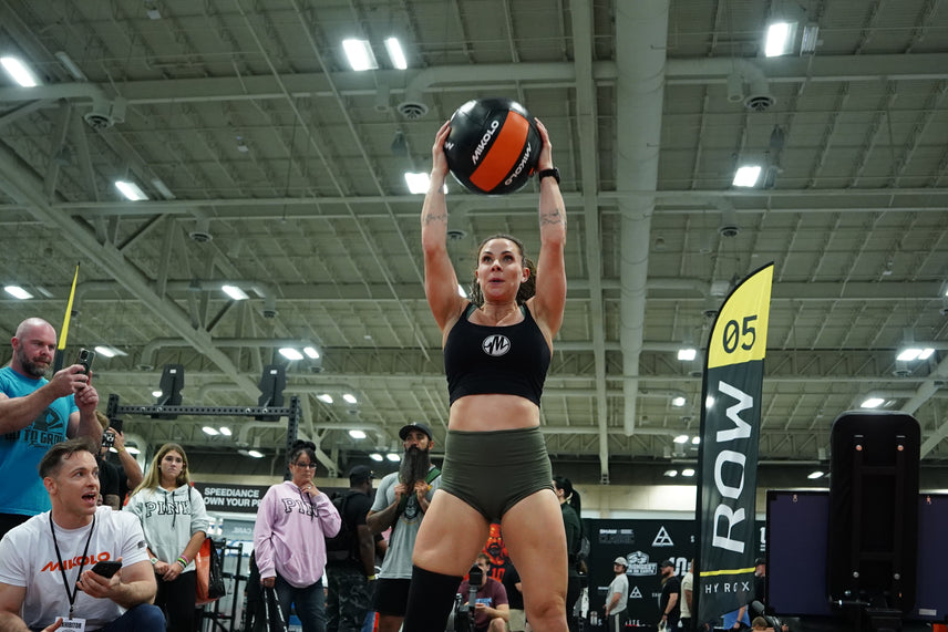 Female athlete holding a black and orange medicine ball overhead at an indoor fitness event with spectators watching