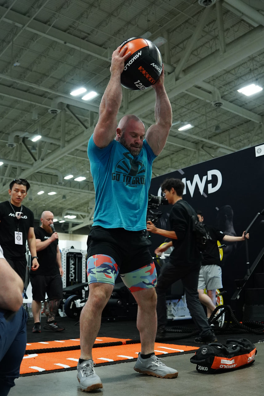 A muscular bearded man in a blue shirt and camouflage shorts holds a black and orange 12kg medicine ball overhead.