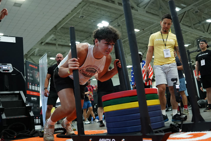 A young man pushes a weighted sled loaded with colorful plates across an orange floor while a man in a yellow shirt watches