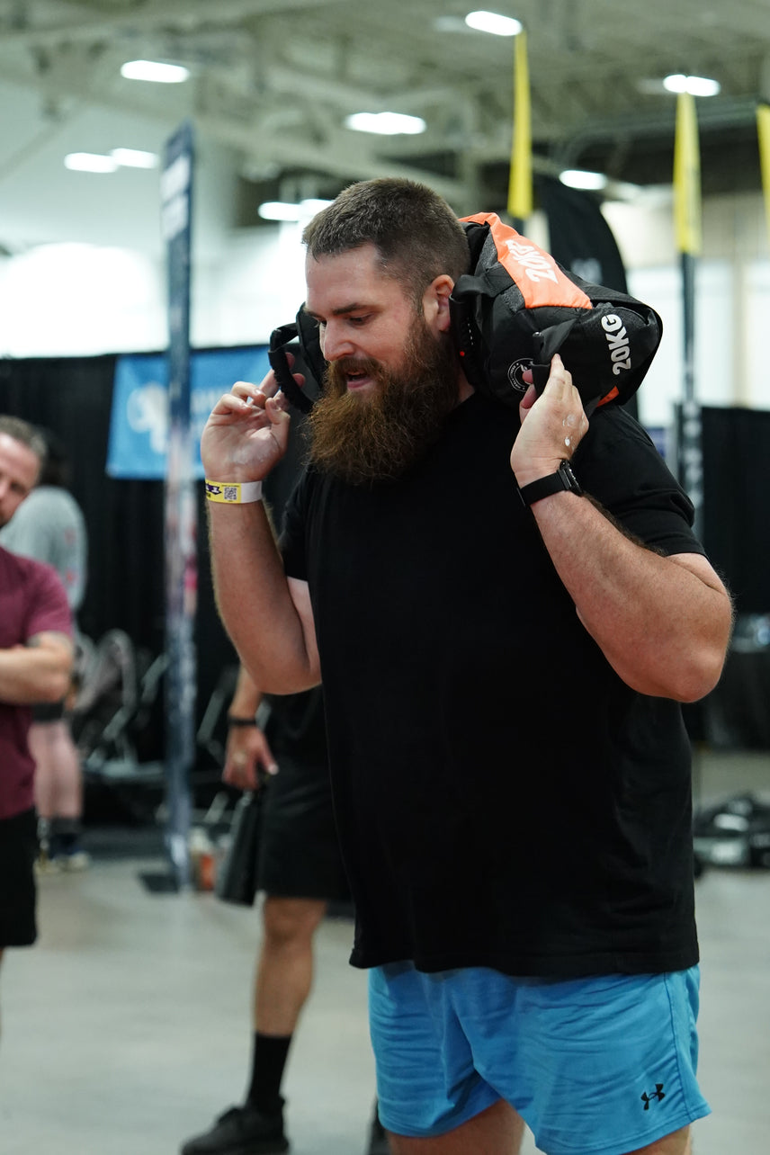 A bearded man in a black t-shirt and blue shorts carries a 20kg black and orange sandbag on his shoulders