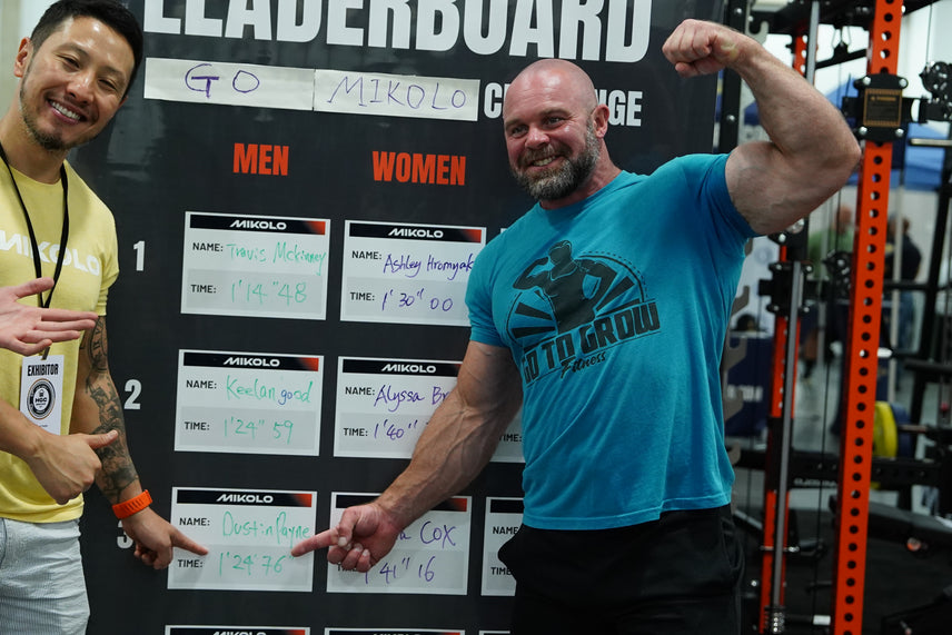 Two smiling men stand before a fitness leaderboard. The man on the left in a yellow shirt points to a name, while the muscula