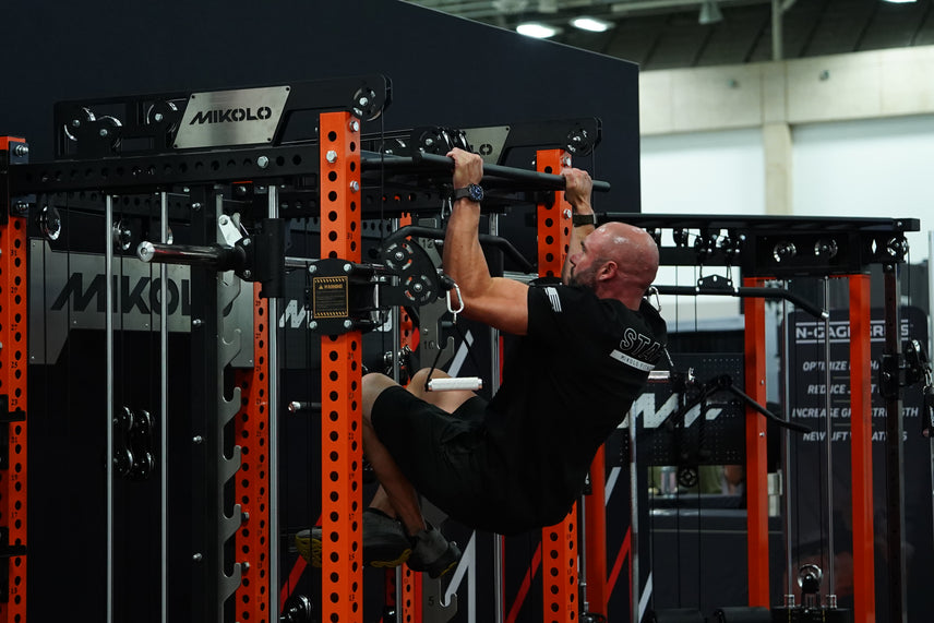 A bald man in black athletic wear performs a hanging leg raise on a black and orange Mikolo power rack