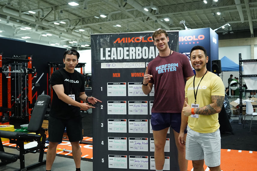 Three men stand in front of a MIKOLO leaderboard at a fitness expo, with the man on the left pointing to the scores