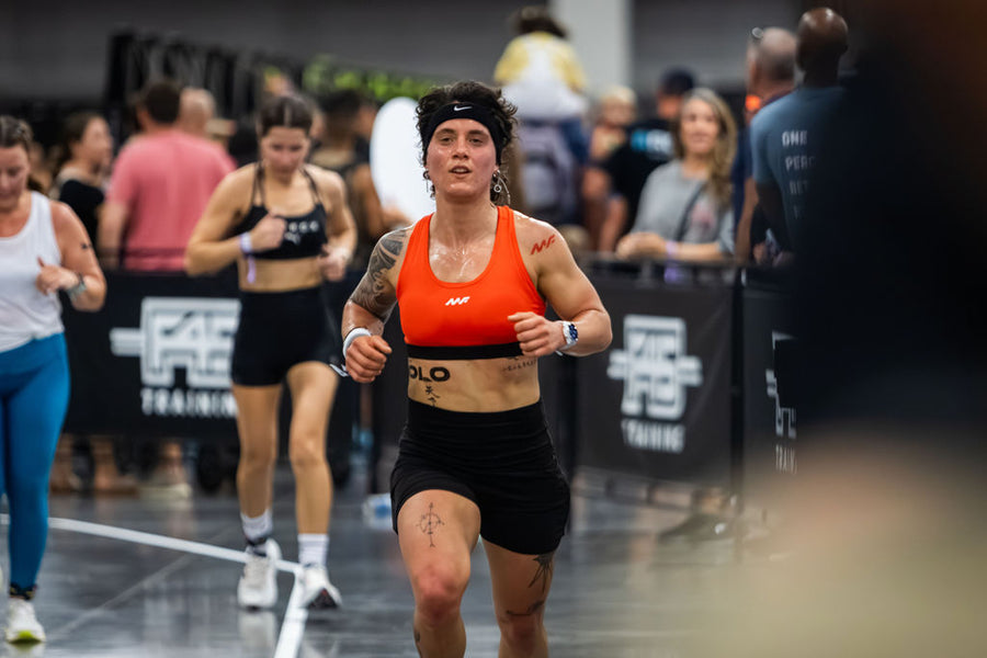 Tattooed woman runs in orange sports bra and black shorts at an indoor fitness event with other participants behind her