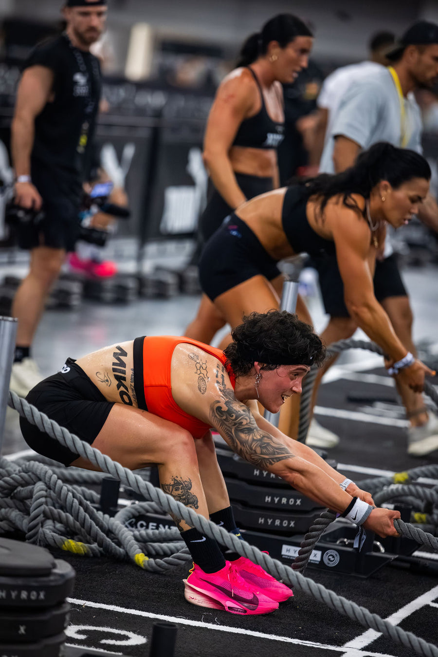 Tattooed woman in orange top and pink shoes pulls a thick rope during a HYROX competition