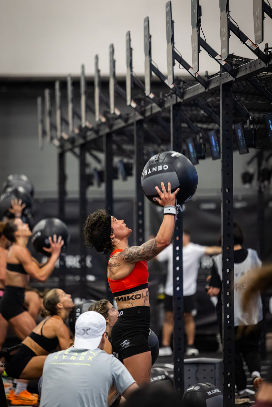 Tattooed woman in red sports bra lifting a black medicine ball overhead in a gym with others exercising in the background