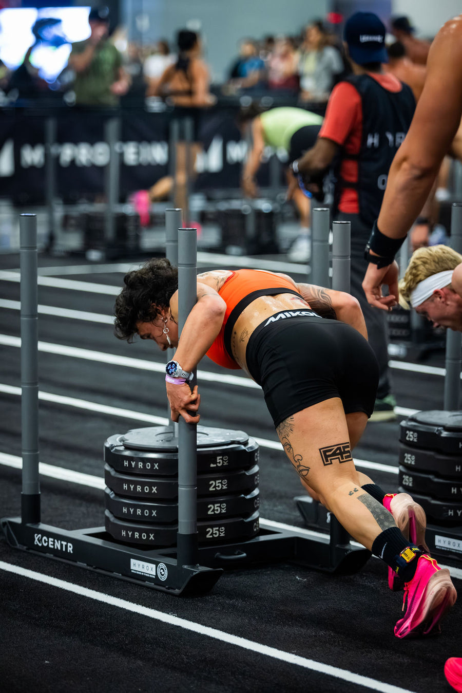 A tattooed woman in an orange sports bra and black shorts pushes a heavy sled loaded with weight plates across a gym floor