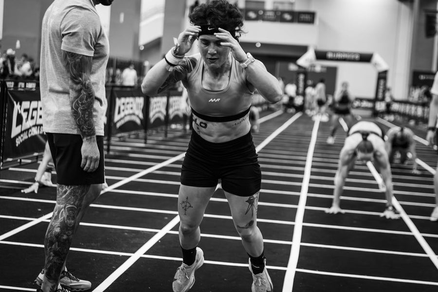 Female athlete with leg tattoos adjusts headband on marked floor next to tattooed man while others exercise in background