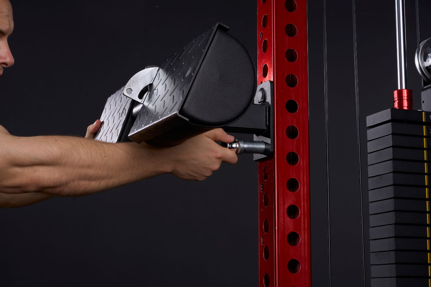 A person inserts a metal pin to secure a black padded gym attachment to a red power rack