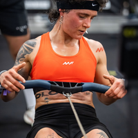 Muscular female athlete with tattoos grips a cable machine handle while wearing an orange sports bra and black shorts