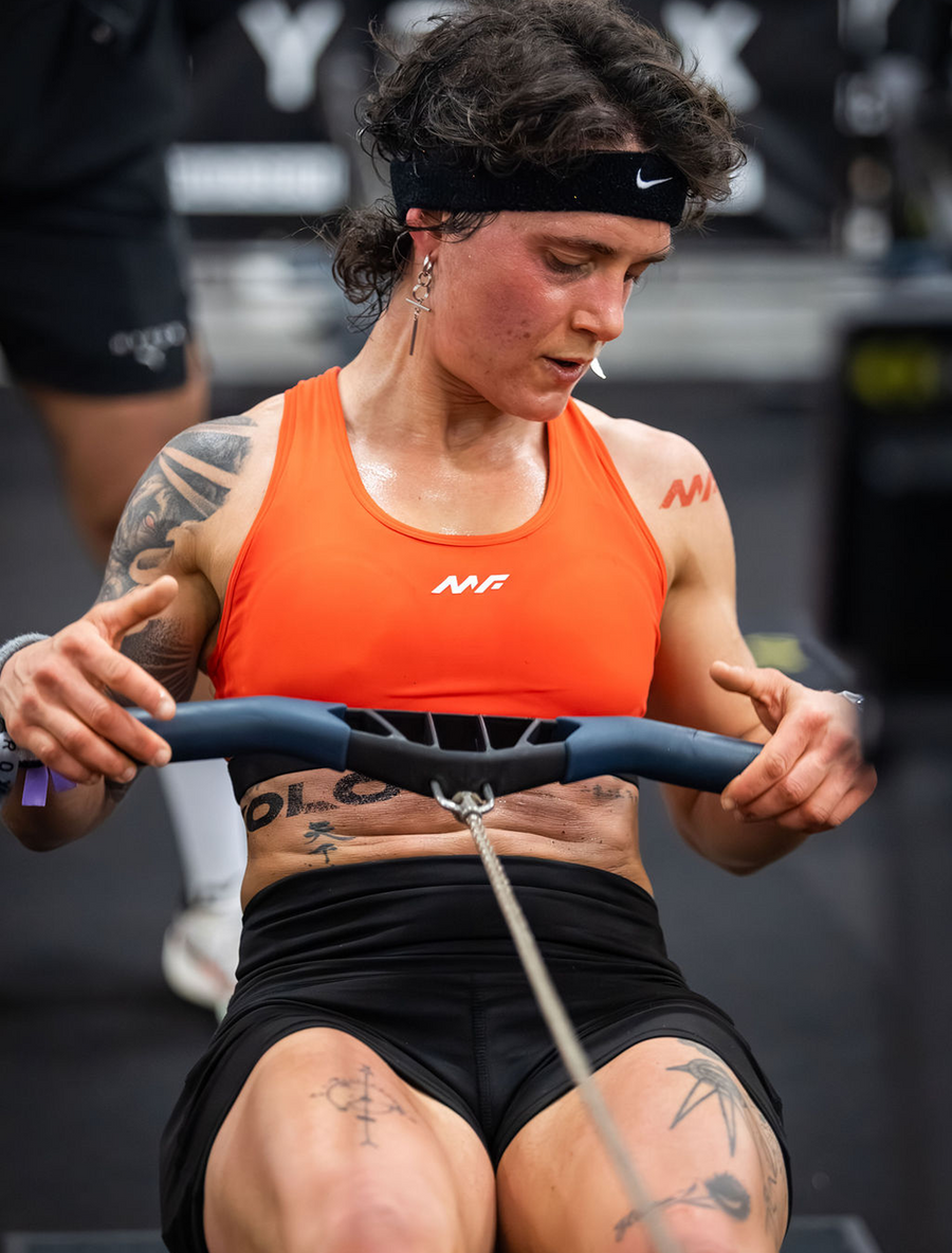Muscular female athlete with tattoos grips a cable machine handle while wearing an orange sports bra and black shorts