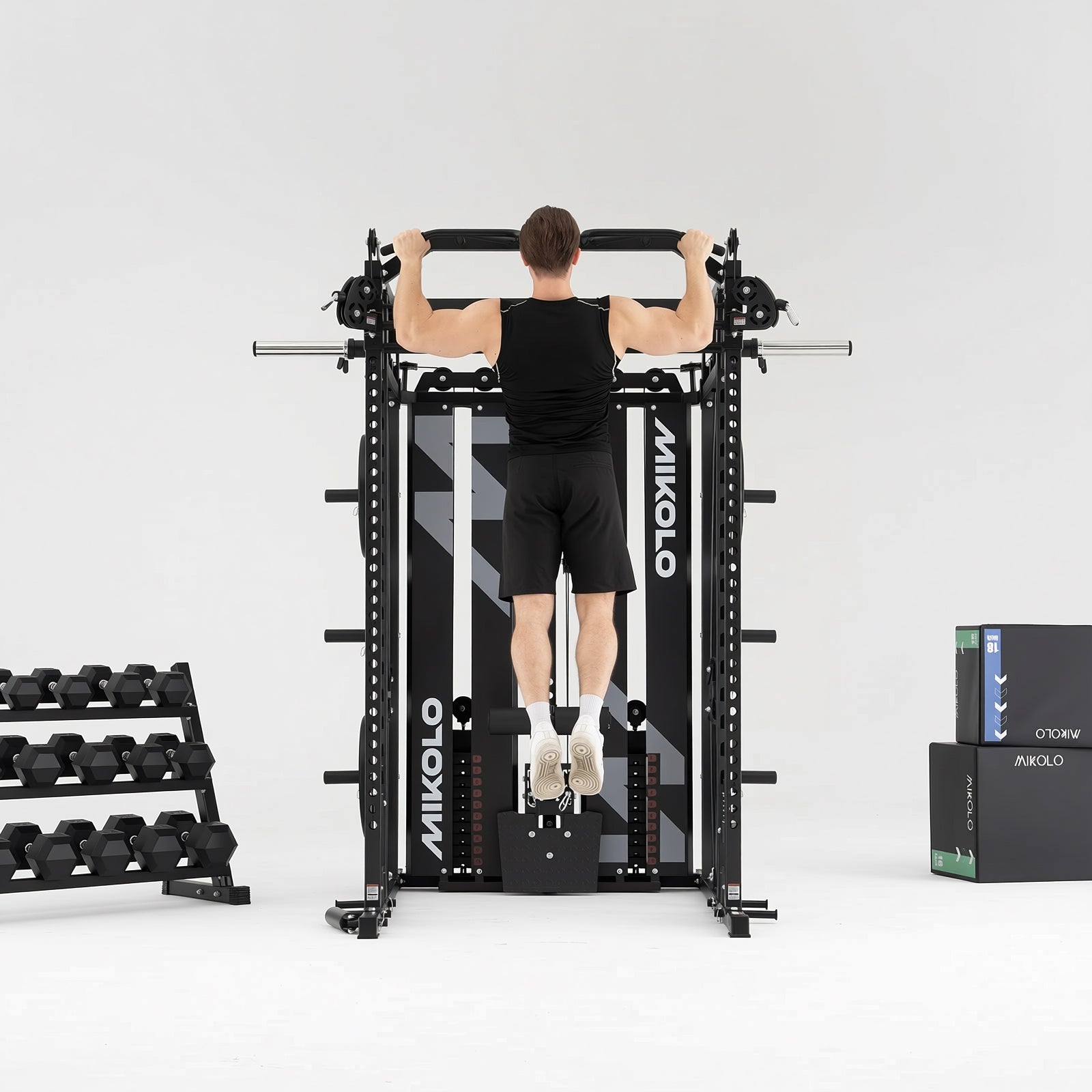 Man doing pull-ups on a black MIKOLO Smith machine, with a dumbbell rack to the left and stacked boxes to the right
