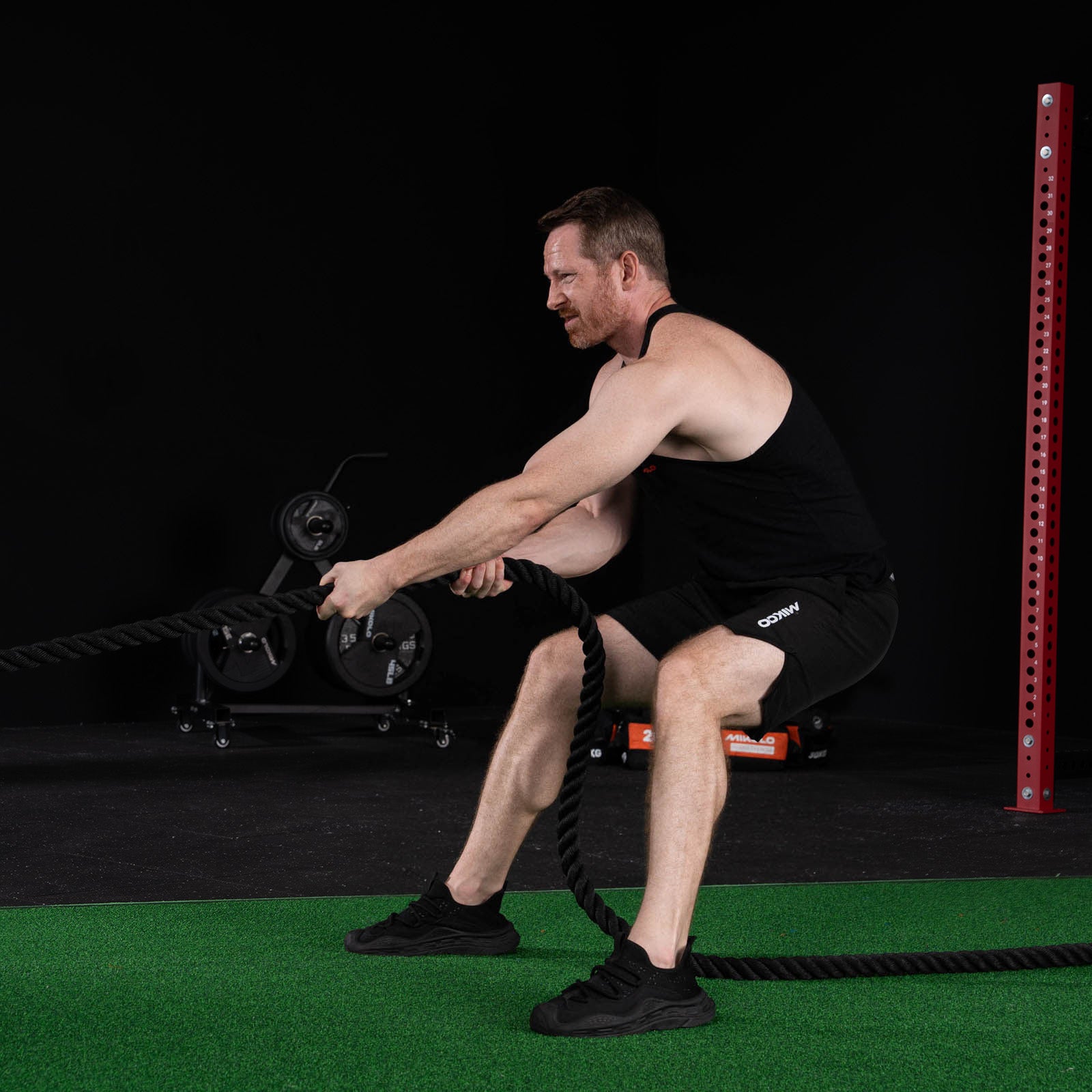 A man in a black tank top and shorts squats while holding thick battle ropes on green gym turf