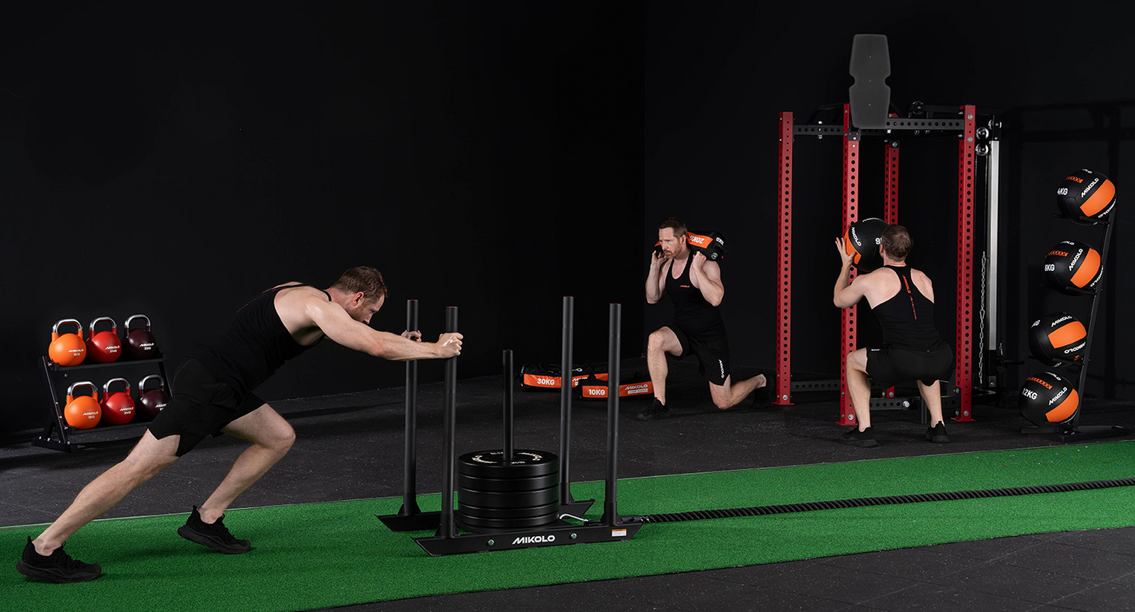 Three men training in a gym pushing a weighted sled, lunging with a sandbag, and squatting with a medicine ball