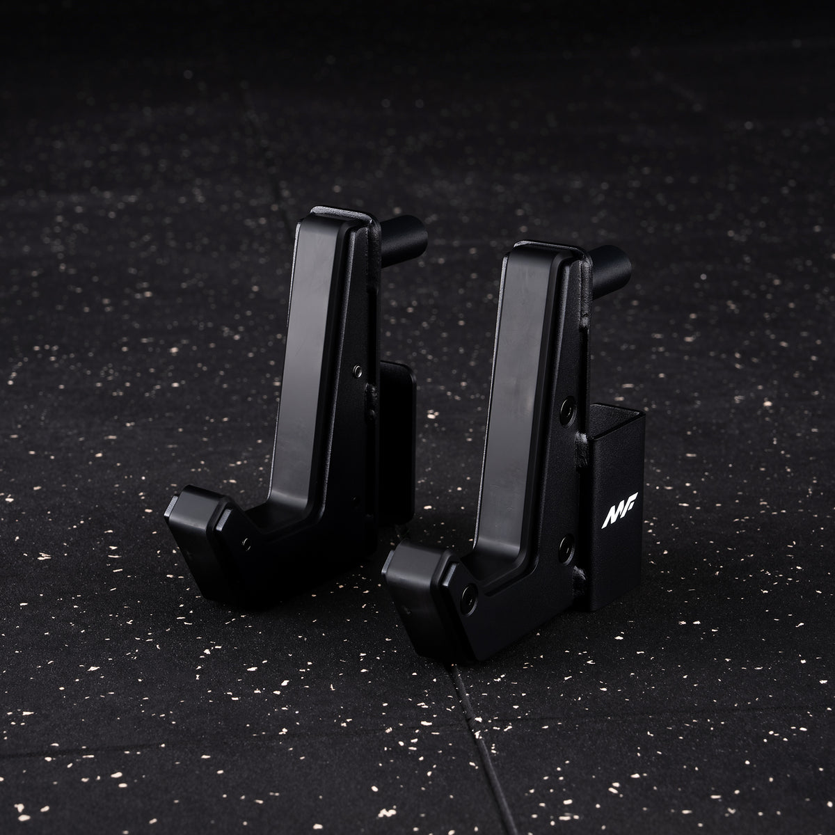 Two black metal J-hooks for a squat rack resting on a speckled gym floor, one displaying a white MF logo
