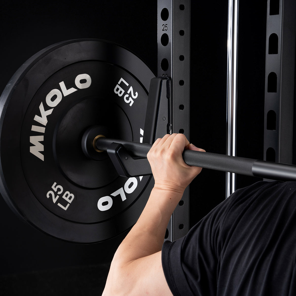 A hand grips a black barbell resting on a J-hook next to a black 25 lb Mikolo weight plate on a squat rack