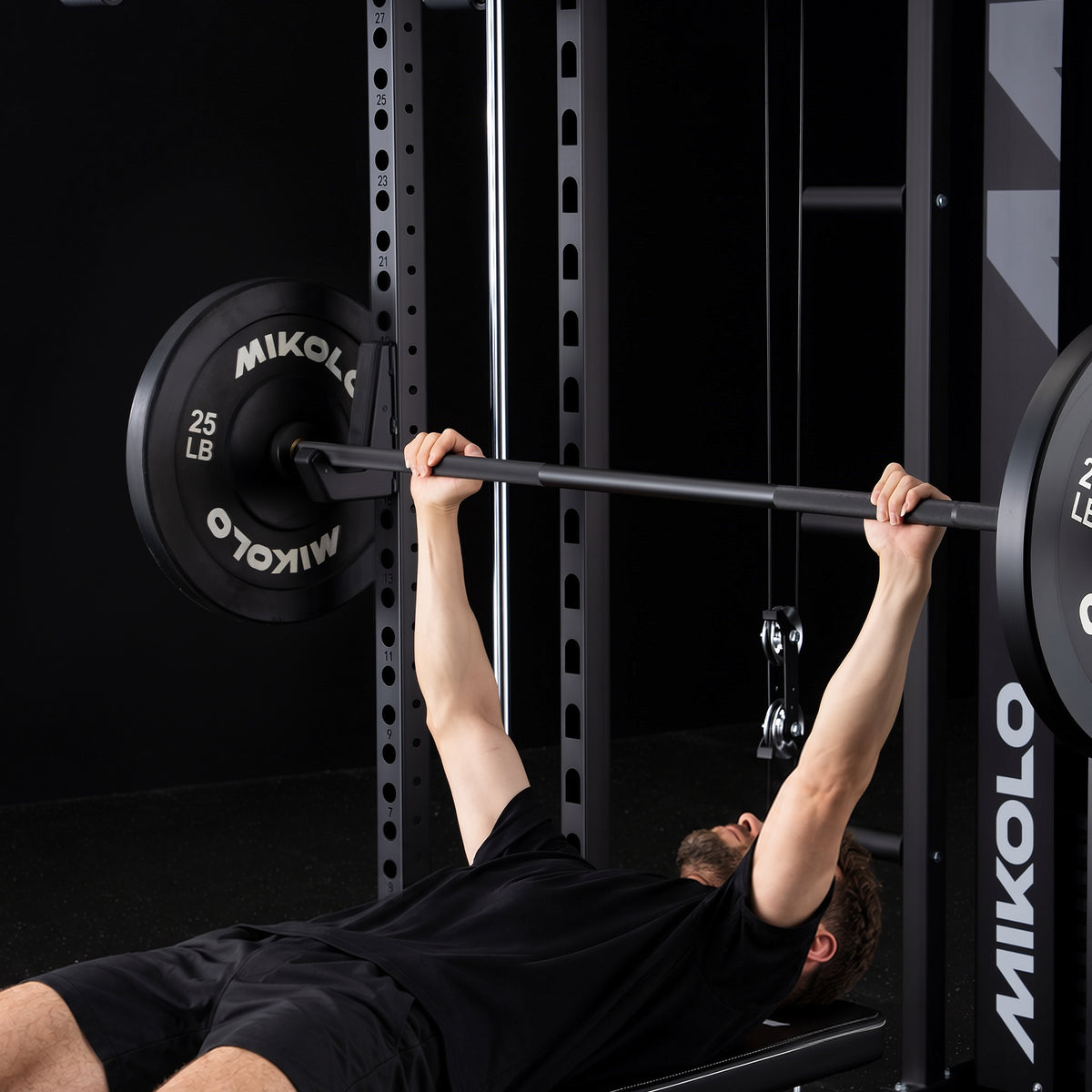 A man lies on a bench pressing a barbell loaded with 25 lb Mikolo weight plates supported by J-hooks on a black rack