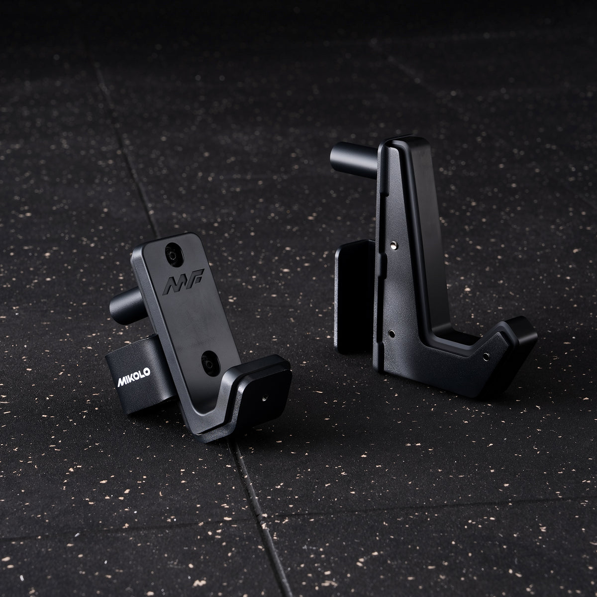 Two black metal MIKOLO J-hooks, one lying flat with an MF logo and one standing upright on a speckled gym floor