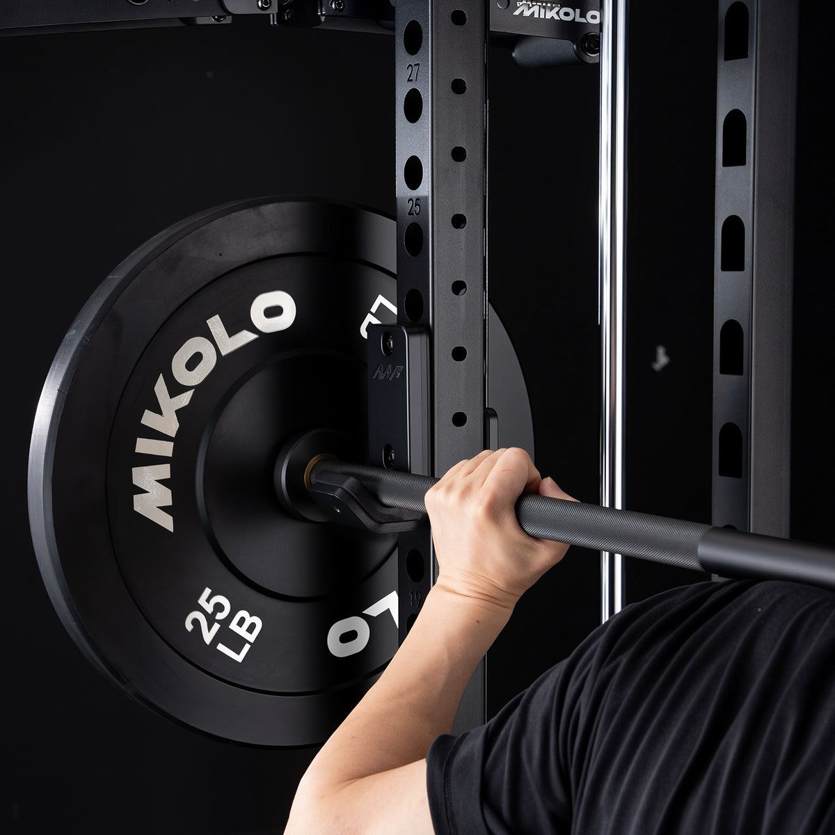 A close-up of a hand gripping a barbell resting on a black squat rack J-hook next to a 25 lb Mikolo weight plate