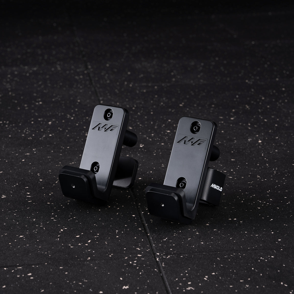 Pair of black metal J-hooks with MF logos and rubber guards sitting on a speckled gym floor, one labeled Mikolo