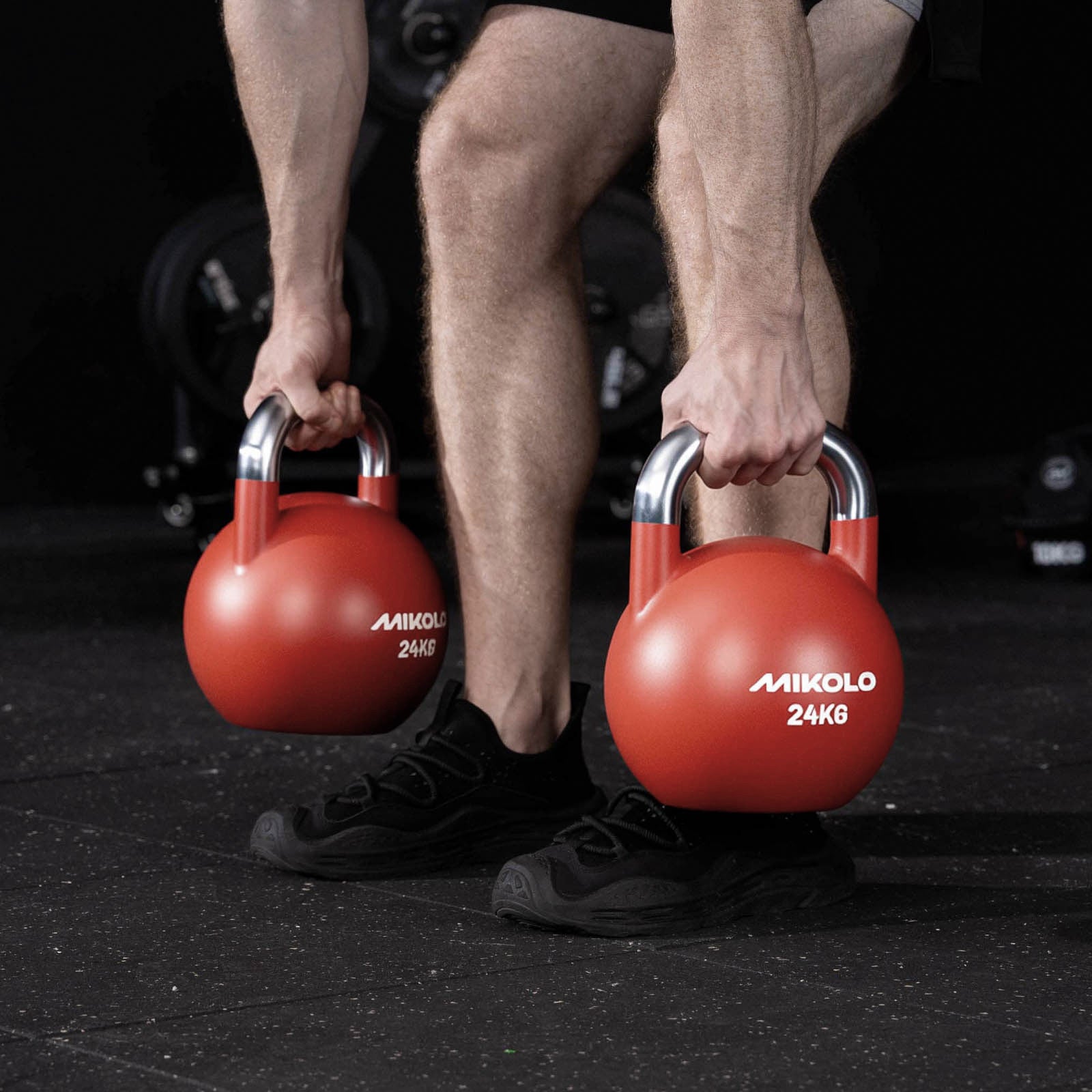 Person holding two red MIKOLO 24kg kettlebells with silver handles while standing on a gym floor wearing black shoes