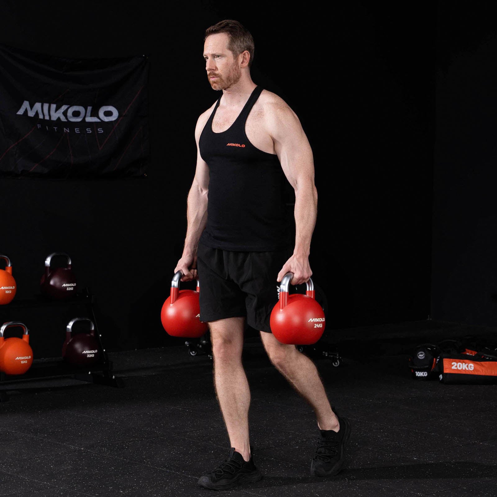 Muscular man in black tank top and shorts holding two red 24kg Mikolo kettlebells at his sides in a gym