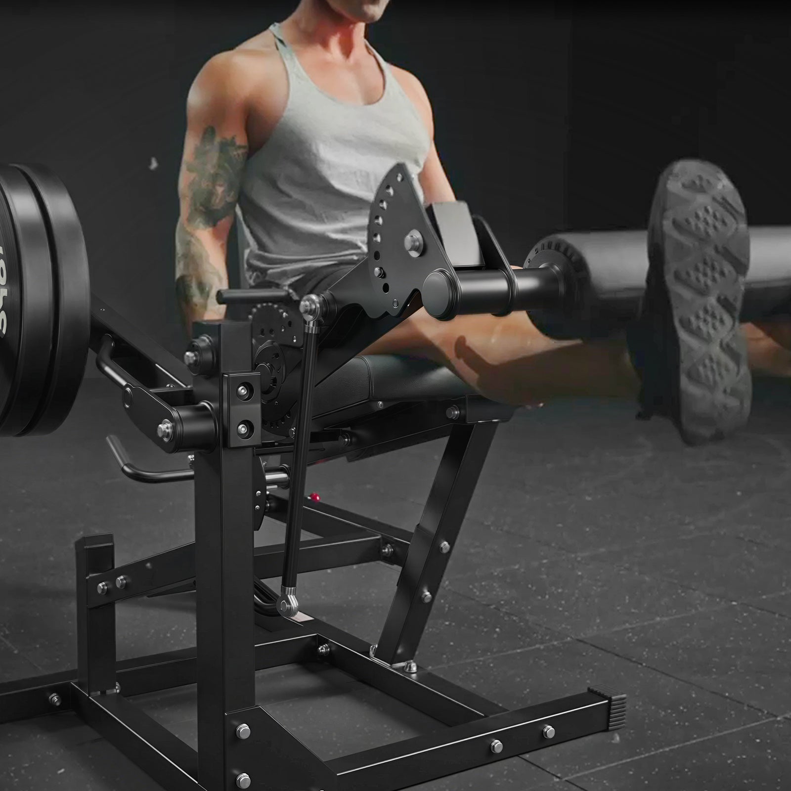A muscular man in a grey tank top performs leg extensions on a black gym machine with weight plates loaded on the side