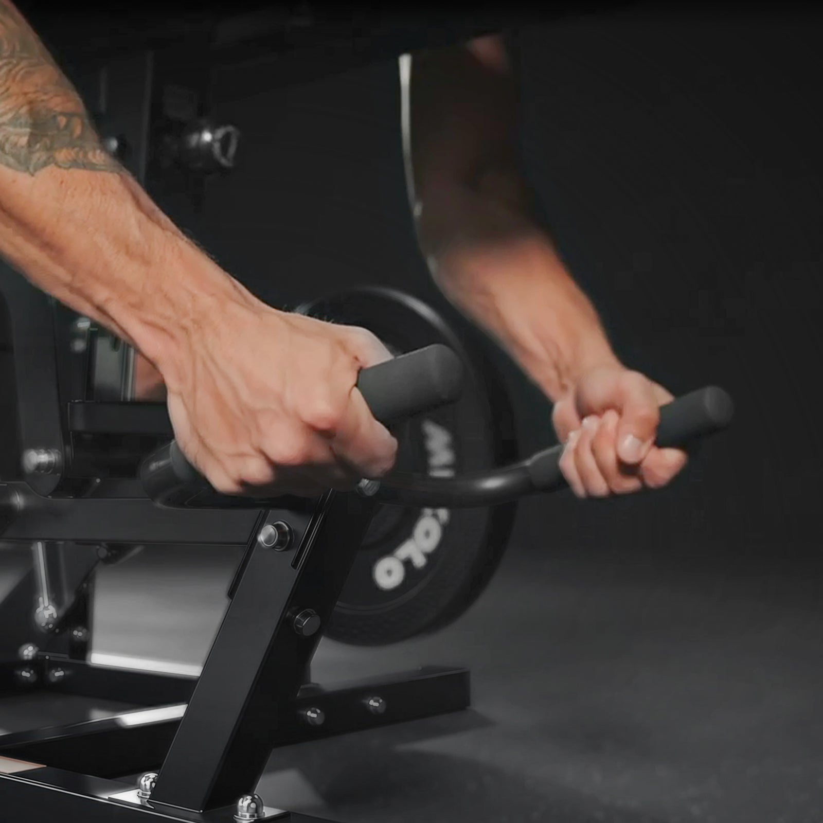 Muscular arms gripping the black handles of a leg curl machine with a weight plate visible in the background