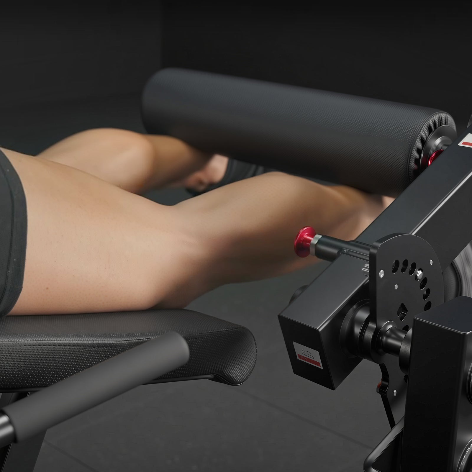Person's legs resting on a black padded bench under a large cylindrical roller of a leg curl machine with red adjustment knob