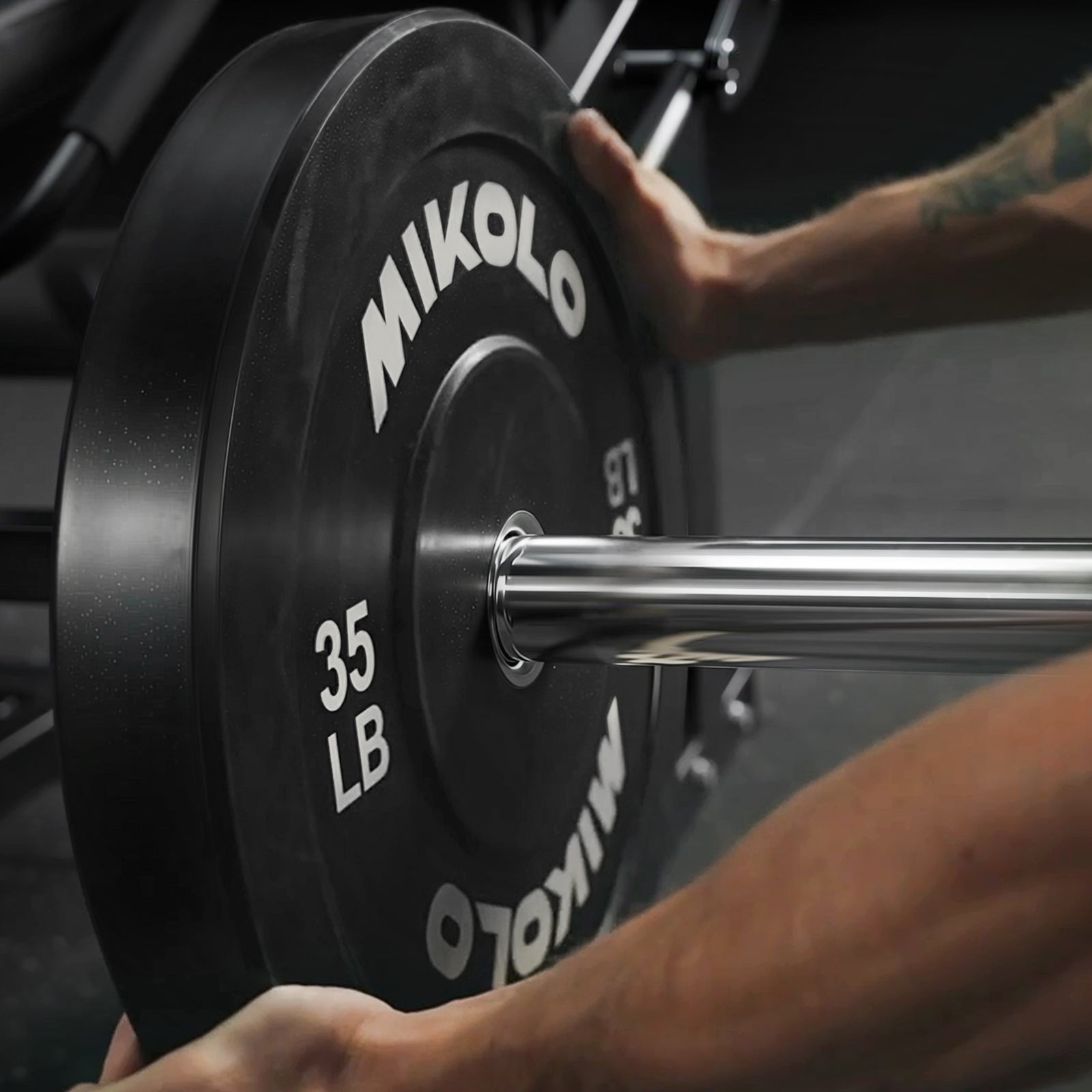 Close-up of hands loading a black Mikolo 35 LB weight plate onto a chrome barbell